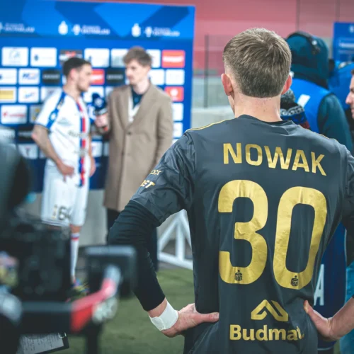 Zawodnik w koszulce z nazwiskiem "NOWAK" i numerem 30 obserwuje wywiad po meczu, w tle widoczni są inni piłkarze oraz dziennikarz.