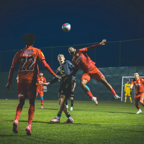 Na zdjęciu widać zawodników w czerwonych strojach podczas meczu piłki nożnej, gdzie jeden z graczy skacze, aby interweniować przy wyskakującym piłce.