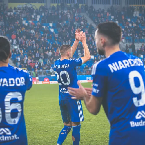 Na zdjęciu widać piłkarzy w niebieskich strojach, którzy klaskają i żegnają się z kibicami po meczu.
