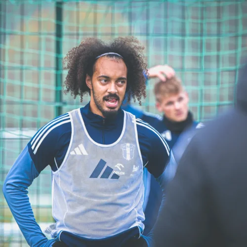 Na zdjęciu widać piłkarza w trakcie treningu, który rozmawia z innymi zawodnikami, a w tle widoczne są bramki do piłki nożnej.