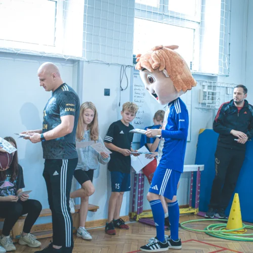 Na zdjęciu widać dzieci i dorosłych w sali sportowej, gdzie jedna z osób przebrana jest w kostium maskotki z dużą głową, a reszta uczestników trzyma kartki i przygląda się wydarzeniu.