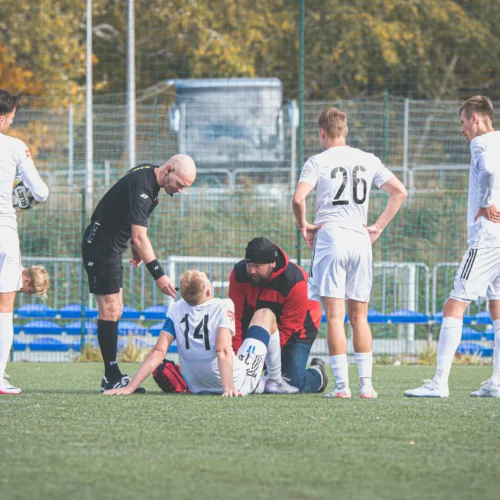 Na boisku piłkarskim zawodnicy zbierają się wokół kontuzjowanego gracza, któremu udzielana jest pomoc medyczna.