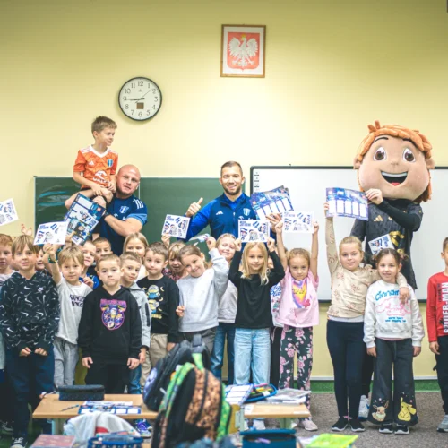 Na zdjęciu znajduje się grupa dzieci w sali lekcyjnej, które trzymają w rękach dyplomy, a wśród nich są też dorośli oraz postać maskotki.