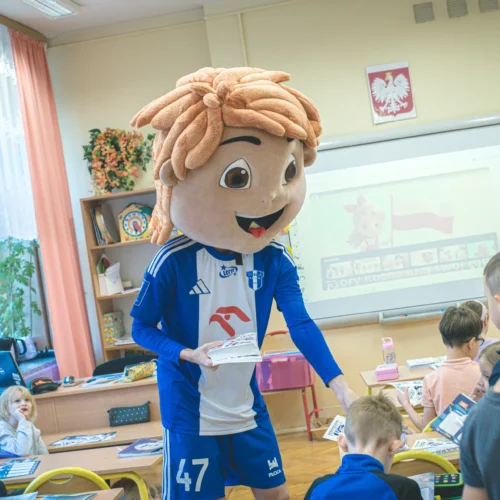 Na zdjęciu widoczna jest osoba w kostiumie maskotki z dużą głową, która rozdaje materiały edukacyjne dzieciom w klasie.