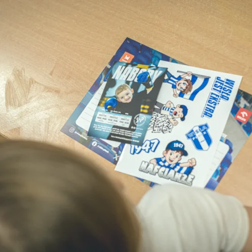 Na zdjęciu znajduje się stół z kolorowymi materiałami promocyjnymi, takimi jak naklejki i ulotki związane z drużyną sportową, na którym widoczna jest część głowy dziecka.