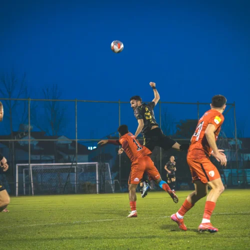 Na zdjęciu widać moment intensywnej akcji piłkarskiej, gdzie gracz w czarno-żółtej koszulce skacze, aby przechwycić piłkę, podczas gdy przeciwnik w pomarańczowym stara się go zablokować.