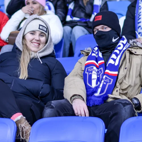 Młoda para kibiców siedzi na trybunach, ubrana w ciepłe kurtki i szaliki drużyny piłkarskiej, uśmiechając się do kamery na tle innych widzów.