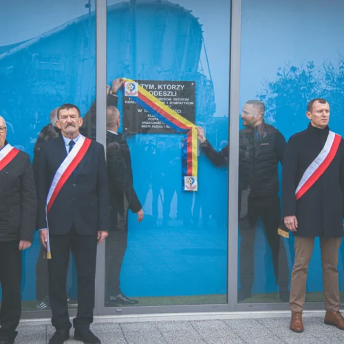 Na zdjęciu widoczna jest ceremonia odsłonięcia tablicy upamiętniającej osoby, które odeszły, z udziałem mężczyzn w strojach galowych, trzymających flagi i wstęgi.