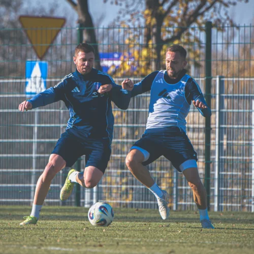 Dwóch piłkarzy w niebieskich strojach intensywnie trenuje na boisku, walcząc o piłkę w słoneczny dzień.