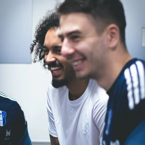 Na zdjęciu trzej mężczyźni w sportowych koszulkach uśmiechają się i rozmawiają w przyjemnej atmosferze.