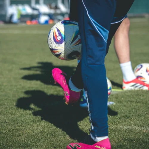 Piłkarz trenuje na boisku, wykonując zwód z piłką w kolorowych butach sportowych.