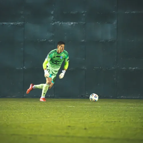 Na zdjęciu widać bramkarza w zielonym stroju, który biegnie w stronę piłki na boisku.