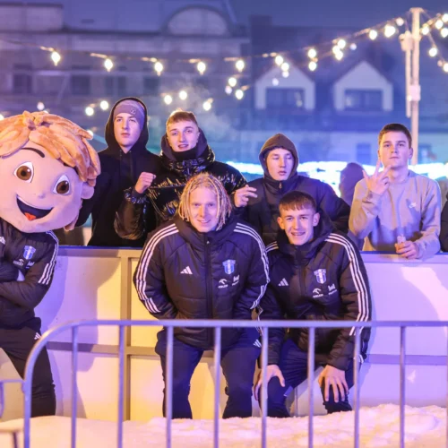 Grupa młodych ludzi, w tym osoba w przebraniu maskotki, pozuje wśród świątecznych dekoracji.