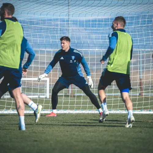 Na zdjęciu widoczna jest grupa piłkarzy trenujących na boisku, z bramkarzem w akcji w pobliżu bramki.