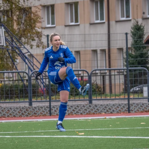 Na zdjęciu widać dwie piłkarki podczas meczu na stadionie, jedna z nich w niebieskim stroju wykonuje szeroki krok, a druga w pomarańczowym czeka na akcję.