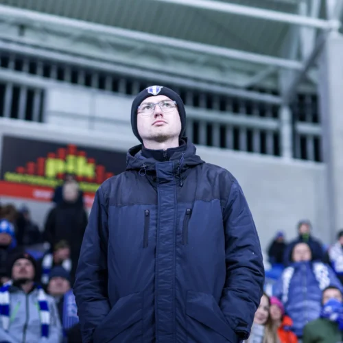 Na zdjęciu widać mężczyznę w ciemnej kurtce i czapce, stojącego na stadionie wśród kibiców.
