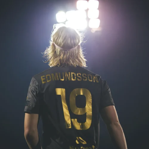 Na zdjęciu widoczna jest sylwetka sportowca w ciemnej koszulce z numerem 19 i nazwiskiem "EDMUNDSSON", stojącego na boisku pod jasnym oświetleniem.