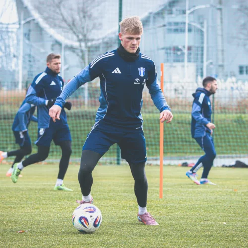 Na zdjęciu widoczny jest piłkarz trenujący na boisku ze złotą piłką, a w tle widać innych zawodników oraz częściowo zasłonięte budynki.