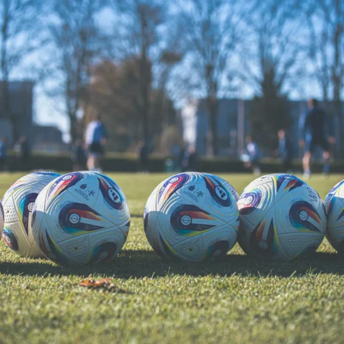 Na zdjęciu widać kilka piłek do futbolu ułożonych na zielonej murawie, z boiskowym tłem i zawodnikami grającymi w oddali.