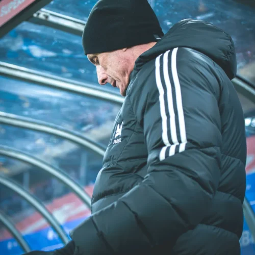 Na zdjęciu widać mężczyznę w czarnym kurtce i czapce, który stoi w boksie trenerskim na stadionie, spoglądając w dół.