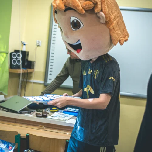 Mężczyzna w przebraniu dużej głowy postaci z kreskówki trzyma kartki, stojąc w pomieszczeniu pełnym ludzi i sprzętu szkolnego.