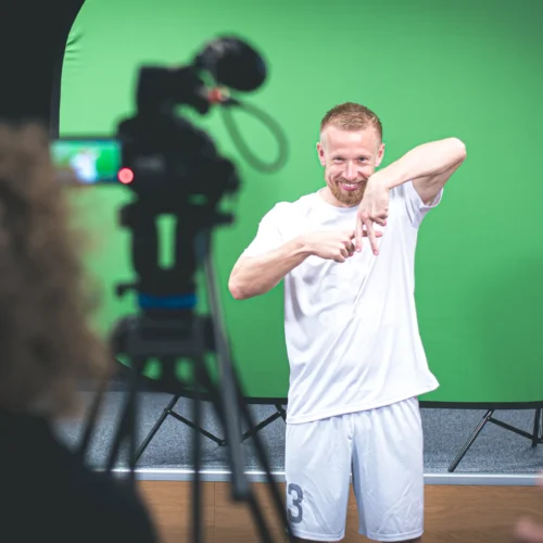 Mężczyzna w białym stroju sportowym pozujący przed kamerą na tle zielonego ekranu.