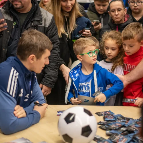 Na zdjęciu widoczny jest zawodnik sportowy, który podpisuje autografy dla grupy dzieci, wśród których jedno w okularach czeka z karteczkami, a na stole leży piłka nożna oraz pakiety z kartami.