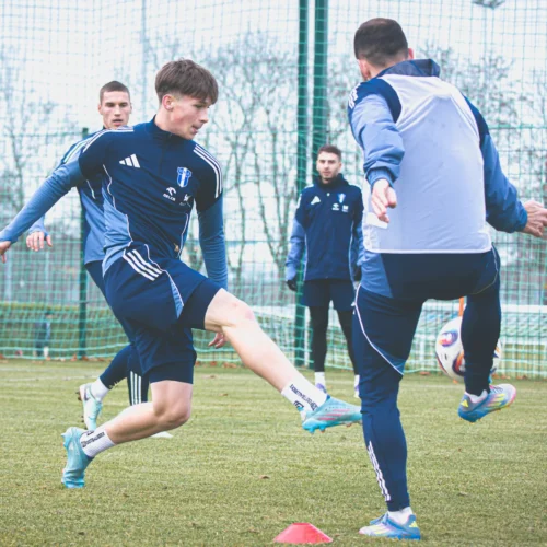 Na zdjęciu zobaczymy grupę młodych piłkarzy trenujących na boisku, podczas gdy jeden z nich wykonuje ćwiczenie, próbując kopnąć piłkę.