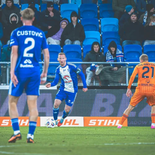 Na zdjęciu widać dwóch piłkarzy w niebieskich strojach podczas meczu, jeden z nich z piłką, a w tle kibice obserwujący rozgrywkę.