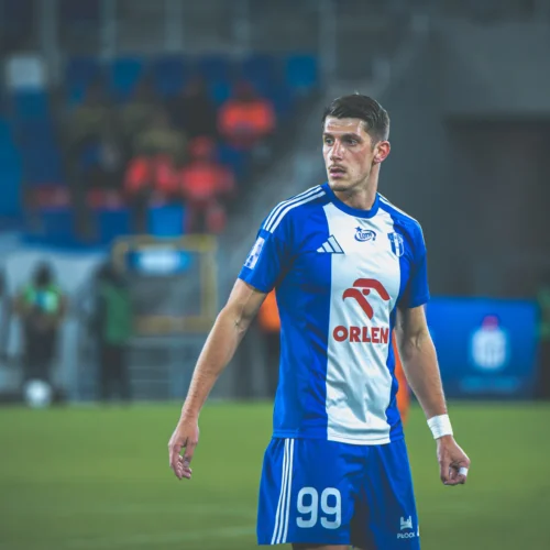 Na zdjęciu widoczny jest piłkarz w niebiesko-białym stroju sportowym, który obserwuje przebieg gry na boisku.