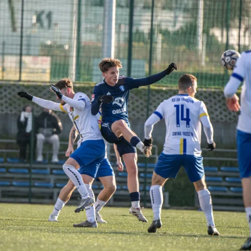Na zdjęciu widać moment dynamicznej akcji podczas meczu piłkarskiego, gdzie zawodnik w niebieskim stroju wykonuje skok z piłką w powietrzu, otoczony przez graczy w białych koszulkach.