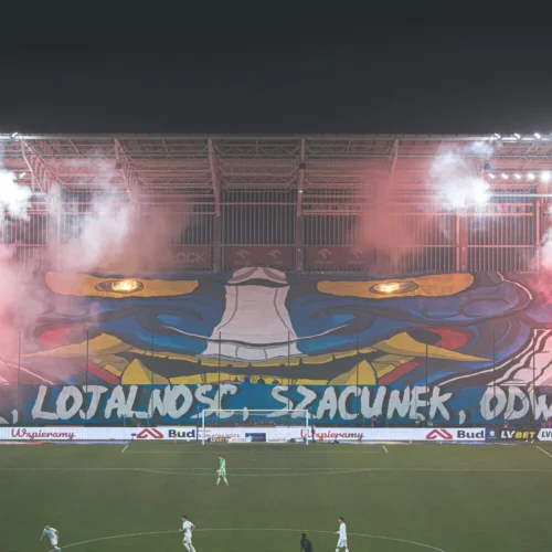 Na zdjęciu widać efektowną oprawę kibiców na stadionie z wielkim muralem i pirotechnika, promującą wartości takie jak honor, lojalność, szacunek i odwaga.