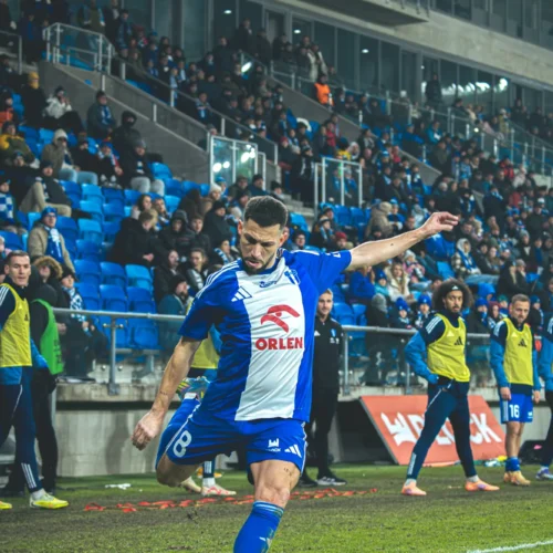 Na zdjęciu przedstawiony jest piłkarz wykonujący rzut rożny na boisku, z widownią w tle.