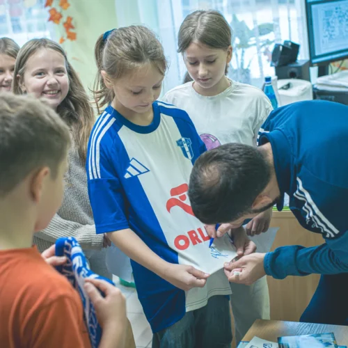 Na zdjęciu grupa dzieci z zainteresowaniem obserwuje dorosłego mężczyznę, który podpisuje koszulkę dla jednego z nich.