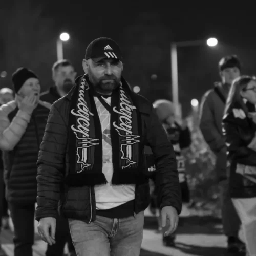 Mężczyzna w czapce i szaliku, ubrany w kurtkę, idzie w tłumie ludzi w nocy.