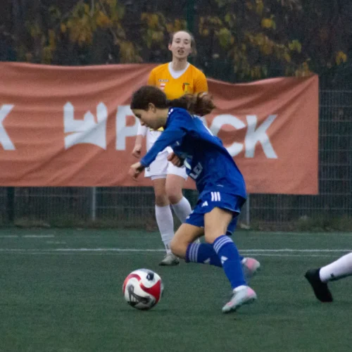 Na zdjęciu widać młode zawodniczki grające w piłkę nożną, z jedną z nich w niebieskim stroju, która schyla się, by zagrać piłkę, podczas gdy druga zawodniczka w żółtej koszulce znajduje się w tle.