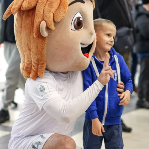 Na zdjęciu widać osobę przebrana za maskotkę sportową, która pozuje z uśmiechniętym chłopcem w niebieskiej bluzie.
