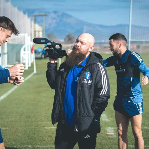 Na zdjęciu widać dwóch piłkarzy w niebieskich strojach oraz mężczyznę z kamerą podczas treningu na boisku.