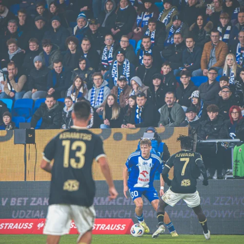 Na zdjęciu przedstawiono scenę ze stadionu podczas meczu piłkarskiego, gdzie zawodnicy rywalizują, a w tle widać licznych kibiców z emocjami na twarzach.