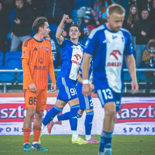 Na zdjęciu widoczny jest piłkarz w niebiesko-białym stroju, który triumfalnie podnosi rękę w górę, podczas gdy jego koledzy z drużyny oraz przeciwnik w pomarańczowym stroju stoją na boisku.