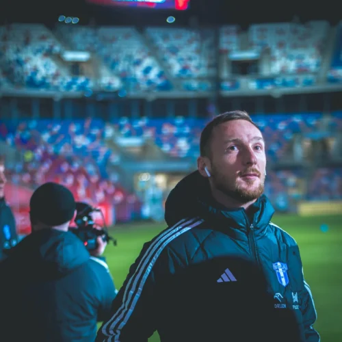 Na zdjęciu widać zawodnika w czarnej kurtce sportowej z białymi detalami, który stoi na stadionie z poważnym wyrazem twarzy, a w tle widoczny jest ekran LED oraz widownia.