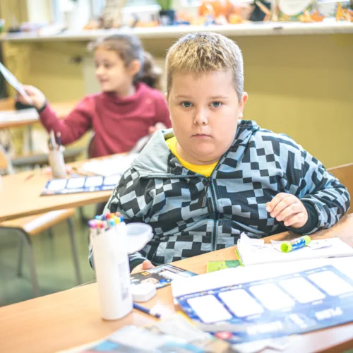 Na zdjęciu widać chłopca w szkolnej klasie, który patrzy w stronę kamery, otoczony przyborami do pisania i rysowania, podczas gdy inna dziewczynka z tyłu odbiera kartkę od nauczyciela.