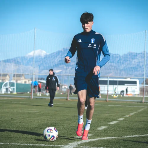Młody piłkarz w stroju sportowym trenuje na boisku, w tle widać góry i innych zawodników.