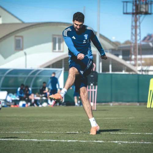 Na zdjęciu widoczny jest piłkarz w niebieskim stroju trenujący na boisku, wykonujący kopnięcie piłki w kierunku bramki.