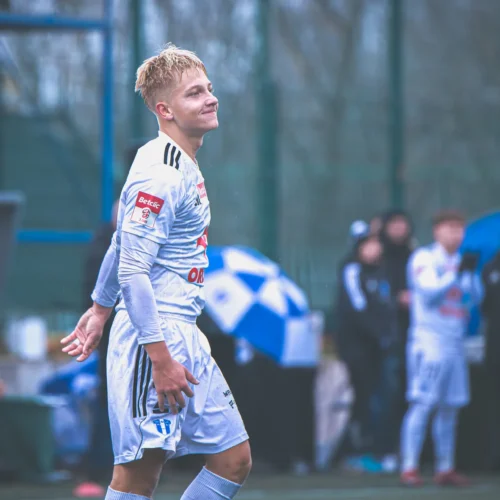 Młody piłkarz w białym stroju z uśmiechem na twarzy, idący po boisku, z widownią w tle.