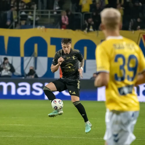 Na zdjęciu dwóch piłkarzy rywalizuje na boisku, a w tle widoczni są kibice z transparentami.