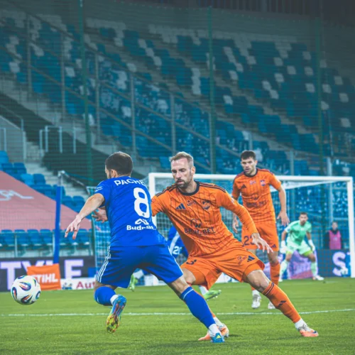 Na zdjęciu widzimy dynamiczną sytuację z meczu piłkarskiego, w której zawodnik w niebieskim trykocie stara się ominąć bramkarza w pomarańczowym stroju.