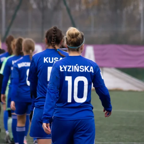 Na zdjęciu widać piłkarki w niebieskich strojach sportowych, idące w kierunku boiska, z widocznymi numerami na plecach.