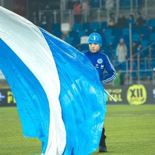 Na zdjęciu widać chłopca ubranym w niebieskie ubranie sportowe, który dzierży dużą, niebiesko-białą flagę na boisku piłkarskim.