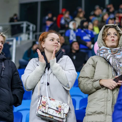 Na zdjęciu widać trzy kobiety w różnych strojach, które są skupione na wydarzeniu na stadionie, z widownią w tle.
